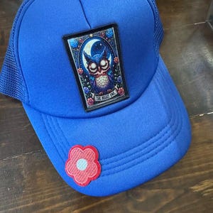 Op de afbeelding: Koningsblauwe truckerpet met een mesh achterkant en een gebogen klep. De pet heeft een patch met een uil-illustratie en de tekst "THE NIGHT OWL". Een kleine roze en rode bloem zit op de klep.