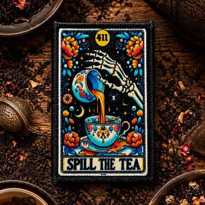 May include: A rectangular embroidered patch featuring a skeleton hand pouring tea from a floral cup into another cup. The design includes vibrant orange and blue flowers, a yellow moon with the number 411, and the text SPILL THE TEA at the bottom.