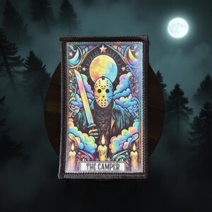 May include: A rectangular patch featuring a masked figure holding a rainbow-colored blade, set against a backdrop of a full moon and colorful clouds. The text "THE CAMPER" is at the bottom. The design has a gothic and horror theme.