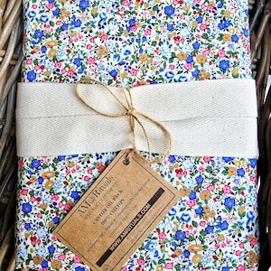 Puede incluir: Una bolsa de tela blanca con un patrón floral en tonos azules, rosas y amarillos. La bolsa está atada con una cinta de cuerda natural y una etiqueta de papel marrón que dice "AM Rituals Naturally Sourced Castor Oil Pack 100% Cotton Handmade in England Organic & Unbleached GOTS Certified Cotton WWW.AMRITUALS.COM".