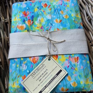 Puede incluir: Un paquete de aceite de ricino rectangular hecho de 100% algodón con un fondo azul vibrante y un patrón floral colorido de flores naranjas, amarillas y rosas. Está envuelto en una cinta de espiga beige ancha atada con cordel y una etiqueta de papel.