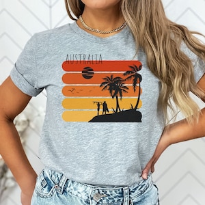 May include: A gray t-shirt with a retro sunset graphic and the word "AUSTRALIA" printed on it. The graphic features palm trees, a surfboard, and two figures standing on a beach.