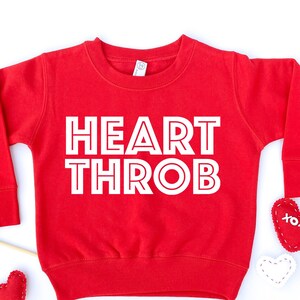 Op de afbeelding: Rode longsleeve trui met de tekst "HEART THROB" in witte omrande letters.