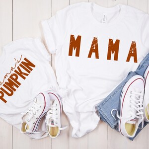 Może przedstawiać: Dwie białe koszulki z pomarańczowym tekstem. Jedna koszulka ma napis "mama", a druga "mama's pumpkin".