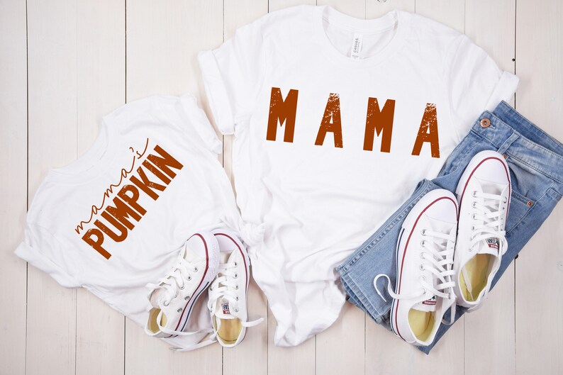 Może przedstawiać: Dwie białe koszulki z pomarańczowym tekstem. Jedna koszulka ma napis "mama", a druga "mama's pumpkin".