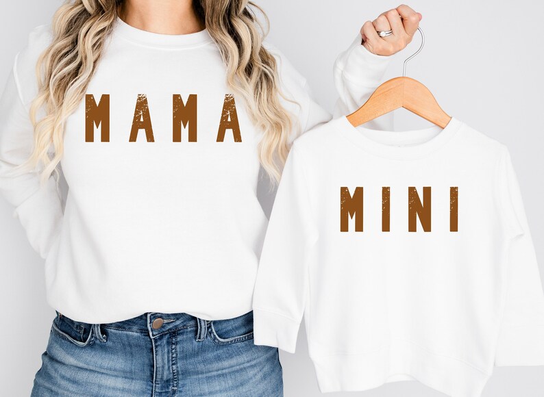 May include: White long-sleeved shirts with brown text. The larger shirt says "MAMA" and the smaller shirt says "MINI".