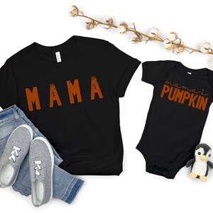 Może przedstawiać: Czarny T-shirt z napisem "MAMA" w pomarańczowych literach i czarny śpioszek dla niemowląt z napisem "mama's PUMPKIN" w pomarańczowych literach.