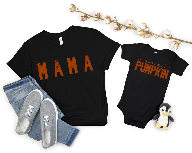 Może przedstawiać: Czarny T-shirt z napisem "MAMA" w pomarańczowych literach i czarny śpioszek dla niemowląt z napisem "mama's PUMPKIN" w pomarańczowych literach.