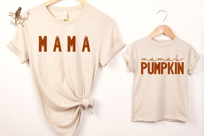 Może przedstawiać: Dwie beżowe koszulki z pomarańczowym nadrukiem. Większa koszulka ma napis "MAMA", a mniejsza "mama's PUMPKIN".