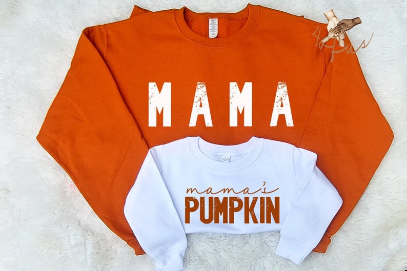 May include: Orange and white matching sweatshirts. The larger sweatshirt says "MAMA" in white with a distressed look. The smaller sweatshirt says "mama's PUMPKIN" in orange with a distressed look.