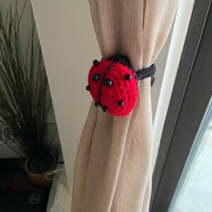 Handmade crochet ladybug curtain holder with black spots and a red shell, tied around a neutral beige linen curtain.