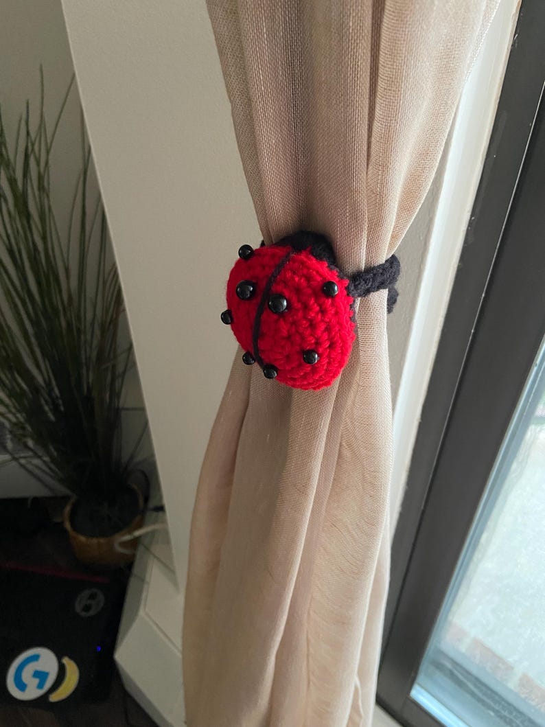 Handmade crochet ladybug curtain holder with black spots and a red shell, tied around a neutral beige linen curtain.