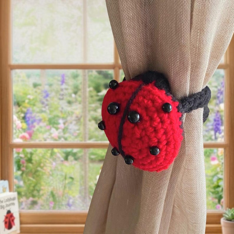 Crochet ladybug curtain tieback displayed on a beige curtain in front of a bright cottage window.