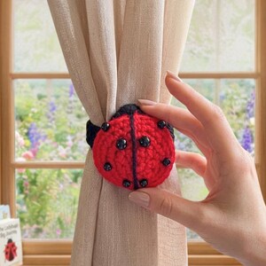 Hand holding a crochet ladybug curtain tieback attached around a beige curtain in front of a window, featuring a red body with black spots and black crochet detailing.