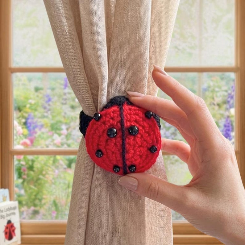 Hand holding a crochet ladybug curtain tieback attached around a beige curtain in front of a window, featuring a red body with black spots and black crochet detailing.