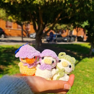 Puede incluir: Tres animales de peluche tejidos a crochet con forma de patos. El pato de la izquierda es amarillo y naranja con un sombrero morado. El pato del medio es blanco con un sombrero morado y una bufanda morada. El pato de la derecha es blanco con un sombrero verde y una bufanda verde.