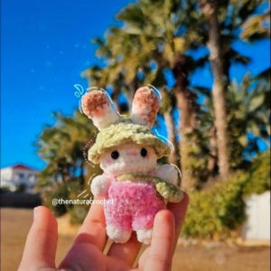 Puede incluir: Una muñeca de conejito hecha a mano con un cuerpo blanco, un peto rosa y un sombrero verde. El conejito tiene orejas marrones y blancas y ojos negros. La muñeca se sostiene en una mano contra un telón de fondo de palmeras y un cielo azul brillante. El texto "@thenaturacrochet" es visible.