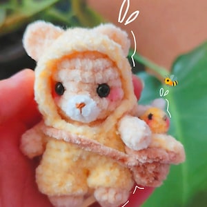 May include: A handmade, crocheted bear wearing a yellow hooded jumpsuit, with a small bee attached. The bear has black eyes, pink cheeks, and a white muzzle. The bear is holding a small bee. The overall color scheme is soft yellow and beige.