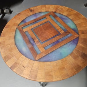 May include: A round table featuring a segmented wooden border and a central square wooden inlay. The surface includes a blue and purple marbled resin inlay surrounding the wood, supported by thin metal hairpin legs.