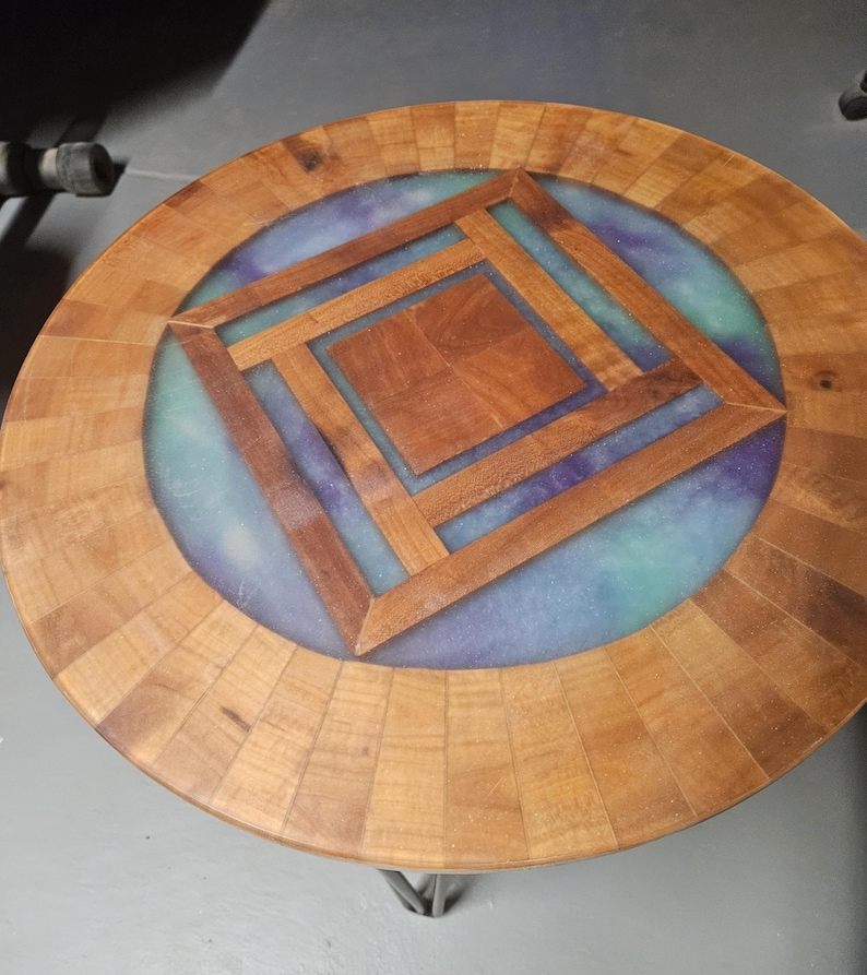 Op de afbeelding: Een ronde tafel met een gesegmenteerde houten rand en een centrale vierkante houten inleg. Het oppervlak bevat een blauw en paars gemarmerde harsinleg rondom het hout, ondersteund door dunne metalen haarspeldpoten.