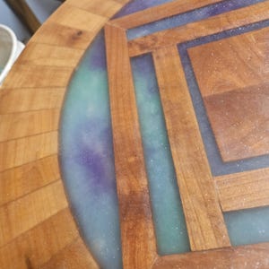 Op de afbeelding: Een ronde houten tafel met een geometrisch inlegpatroon van lichtbruine houtsegmenten gescheiden door een doorschijnende, glinsterende harsvulling in tinten blauwgroen en paars, wat zorgt voor een glad oppervlak.