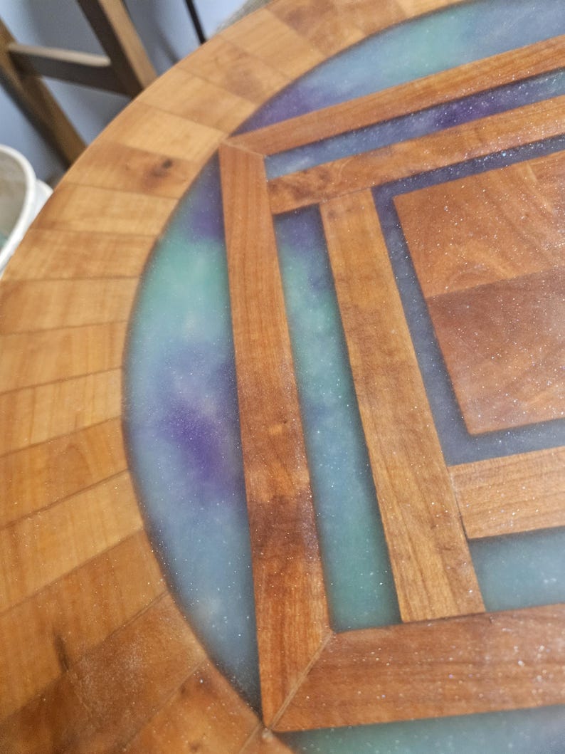 Op de afbeelding: Een ronde houten tafel met een geometrisch inlegpatroon van lichtbruine houtsegmenten gescheiden door een doorschijnende, glinsterende harsvulling in tinten blauwgroen en paars, wat zorgt voor een glad oppervlak.