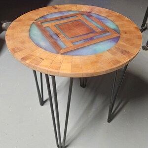 Op de afbeelding: Een ronde bijzettafel met een houten blad met een gesegmenteerde rand en een centrale vierkante geometrische inleg, omringd door een ring van doorschijnende blauwe en paarse hars op vier zwarte metalen haarspeldpoten.