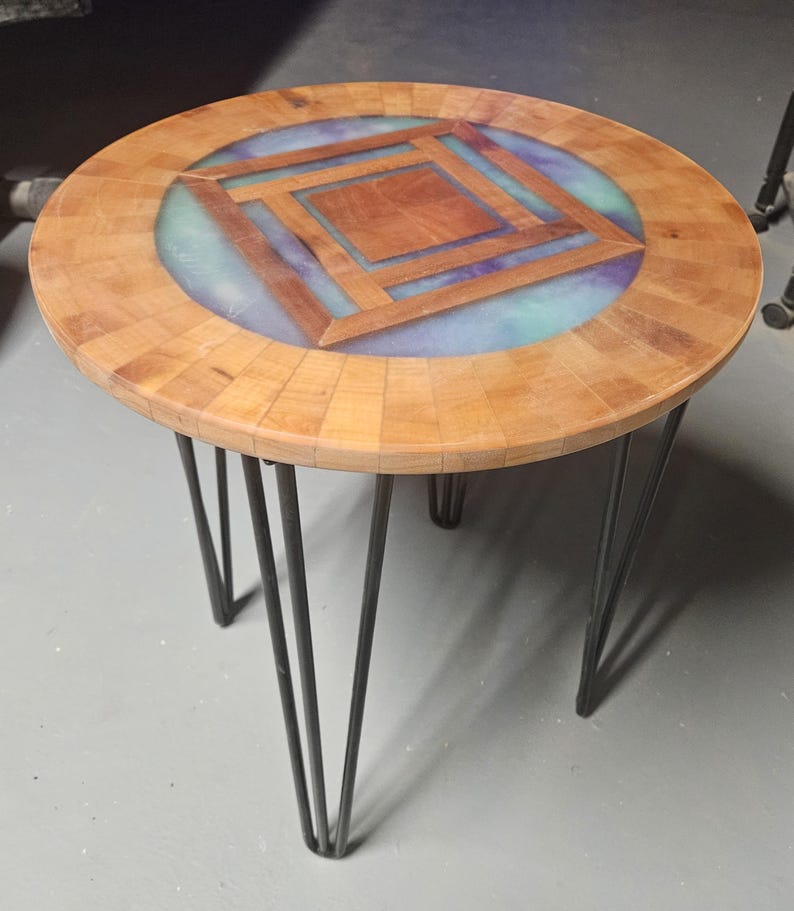 Op de afbeelding: Een ronde bijzettafel met een houten blad met een gesegmenteerde rand en een centrale vierkante geometrische inleg, omringd door een ring van doorschijnende blauwe en paarse hars op vier zwarte metalen haarspeldpoten.