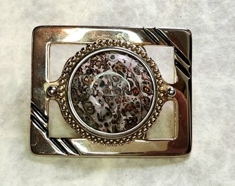May include: A gold-toned metal belt buckle with a square frame and a round, brown and white stone in the center.