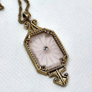 May include: A gold-toned pendant necklace featuring an elongated octagonal frame with a beaded edge. The center contains a pale pink, sunburst-patterned glass insert with a single small, clear, round-cut stone set in the very middle.