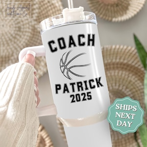 May include: White tumbler with a black basketball graphic and the text "COACH PATRICK 2025". The tumbler has a white handle and a clear lid with a straw. The tumbler is held by a person wearing a white sweater.