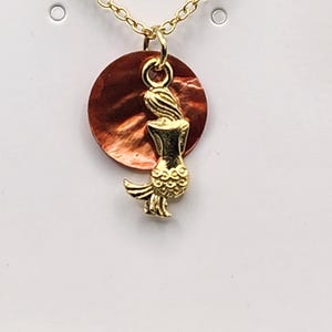 May include: Gold-tone necklace featuring a mermaid pendant. The pendant has a gold mermaid figure in front of a round, reddish-brown shell. The necklace has a delicate chain and is displayed on a white background.