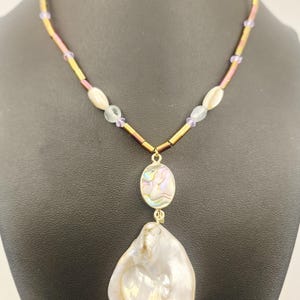 May include: A handcrafted necklace featuring a large, iridescent, pearlescent pendant. The necklace has a gold-toned chain with purple and clear beads. The pendant is a large, irregular shape, and the necklace is displayed on a black mannequin.