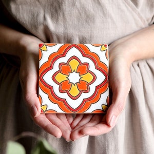 May include: A ceramic tile featuring a geometric pattern in shades of orange, yellow, and white. The tile has a white background with a central flower-like design.
