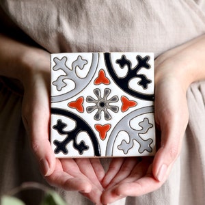 Può includere: Una piastrella di ceramica bianca con un motivo geometrico nero, grigio e arancione. La piastrella presenta un design ripetitivo di fiori e foglie stilizzati.