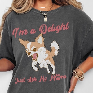 以下が含まれることがあります： ピンクの襟をした漫画のチワワと「I'm a Delight Just Ask My Mom」というピンクの文字が書かれたグレーのオーバーサイズTシャツです。