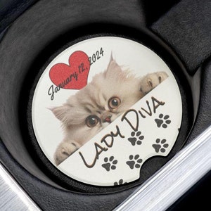 May include: White car cup holder with a round white coaster featuring a white kitten peeking out from behind a red heart with the text "January 12, 2024" and the text "Lady Diva" with black paw prints.