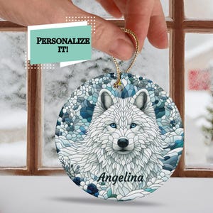 May include: A round ceramic ornament with a stained glass design of a white wolf with blue and green accents. The wolf's name, "Angelina", is written in white lettering at the bottom of the ornament.