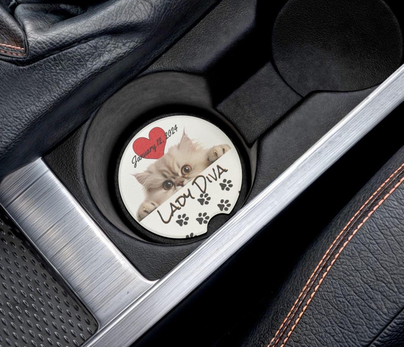 May include: White car cup holder with a round white coaster featuring a white kitten peeking out from behind a red heart with the text "January 12, 2024" and the text "Lady Diva" with black paw prints.