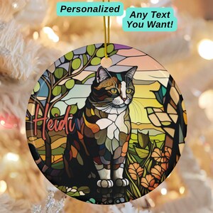May include: A round, personalized cat ornament with a stained-glass effect. The ornament features a calico cat sitting in a garden scene with the name "Heidi" in pink script. Text above reads "Personalized Any Text You Want!" on a light blue background.