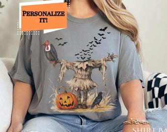 Könnte beinhalten: Graues T-Shirt mit einem Halloween-Motiv. Das Design zeigt eine Vogelscheuche, einen Kürbis, einen Geier, eine Krähe und Fledermäuse. Der Text "PERSONALIZE IT!" befindet sich in einer orangefarbenen Box. Das Shirt ist ein bequemer, lässiger Stil.