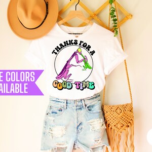 May include: White t-shirt with a graphic of a praying mantis eating a green leaf. The graphic is in a circle with the text "Thanks for a good time" in rainbow colors. The shirt is on a hanger with a straw hat and a woven bag.