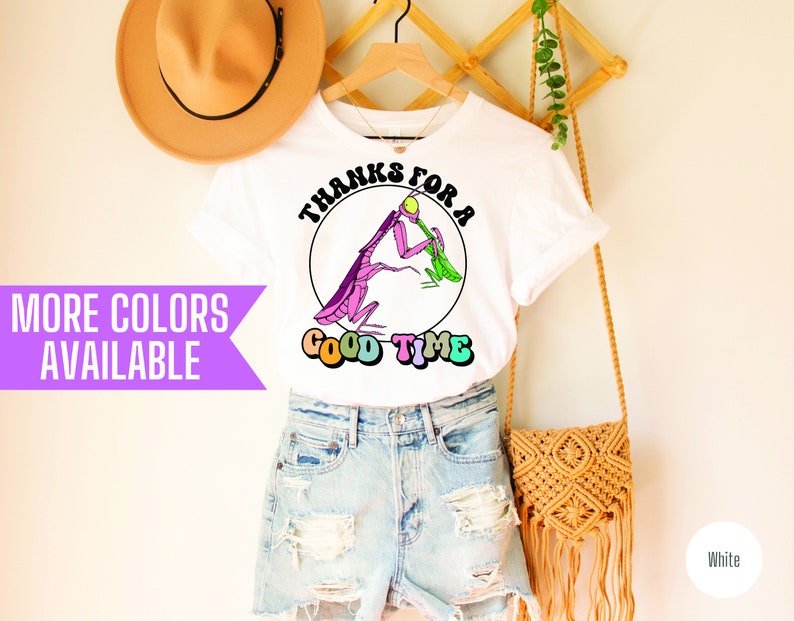 May include: White t-shirt with a graphic of a praying mantis eating a green leaf. The graphic is in a circle with the text "Thanks for a good time" in rainbow colors. The shirt is on a hanger with a straw hat and a woven bag.