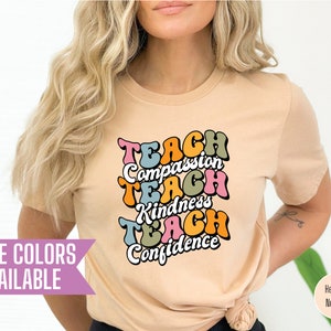 May include: A light beige t-shirt with a graphic design that says "Teach Compassion Teach Kindness Teach Confidence" in a colorful retro font.