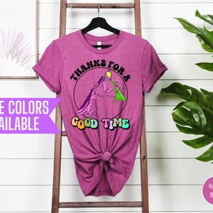 May include: A heather mauve t-shirt with a graphic of a praying mantis holding a green leaf and a yellow ball. The text "Thanks for a Good Time" is printed in a rainbow font around the mantis.