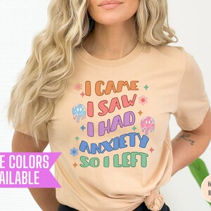 May include: A heather neutral t-shirt with the text "I CAME I SAW I HAD ANXIETY SO I LEFT" in colorful, retro-style lettering. The design includes flowers and melting smiley faces. The shirt is available in more colors.