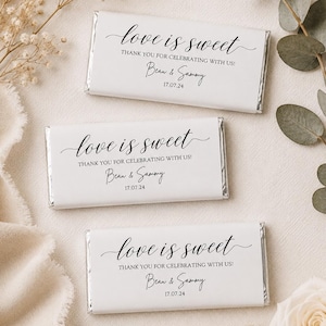 May include: Three rectangular chocolate bars wrapped in white paper with black text that reads love is sweet, THANK YOU FOR CELEBRATING WITH US!, Beau & Sammy, and 17.07.24. The wrappers have silver foil edges visible on the sides of each bar.