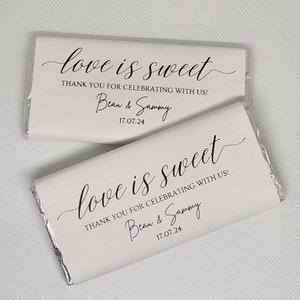 May include: Two rectangular chocolate bars wrapped in white paper with black text. The text reads "love is sweet", "THANK YOU FOR CELEBRATING WITH US!", "Beau & Sammy", and the date "17.07.24". The wrappers have silver foil ends visible.