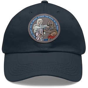 Peut inclure: Une casquette de baseball bleu marine avec un patch circulaire représentant une femme frappant un diable au visage. Le patch porte le texte "Hail Mary Full Of Grace, Punch The Devil In The Face".