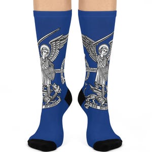 May include: A pair of blue socks with a white graphic of an angel with a sword and shield. The angel is standing on a dragon. The text "Non Serviam" is written on the socks.